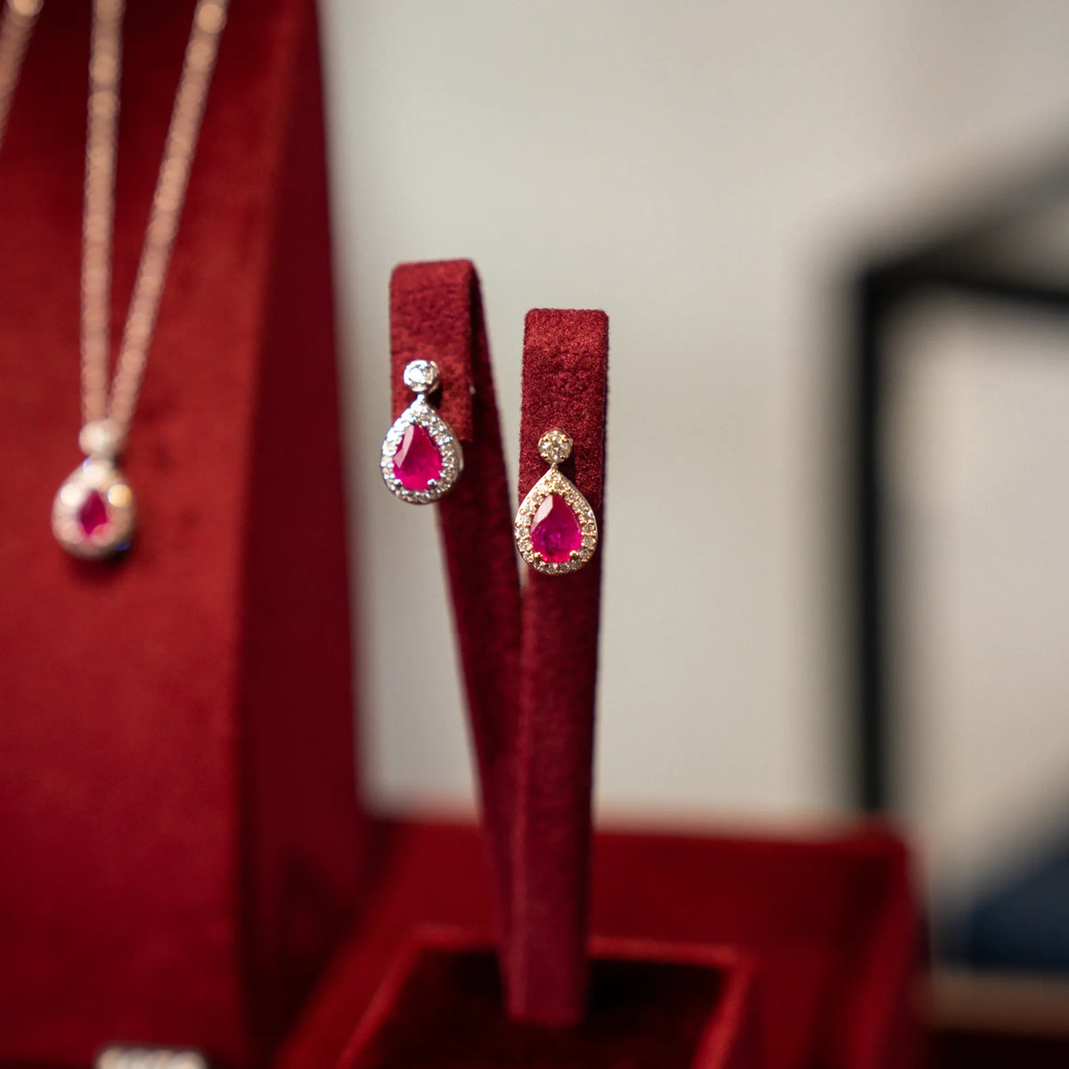 Produktfotografie der weißgoldenen Ohrstecker von Lunatica Roma mit rotem Rubin im Tropfen-Schliff und Diamant-Besatz, während diese auf einem samtigen, weinroten Juwelier-Ständer präsentiert wird