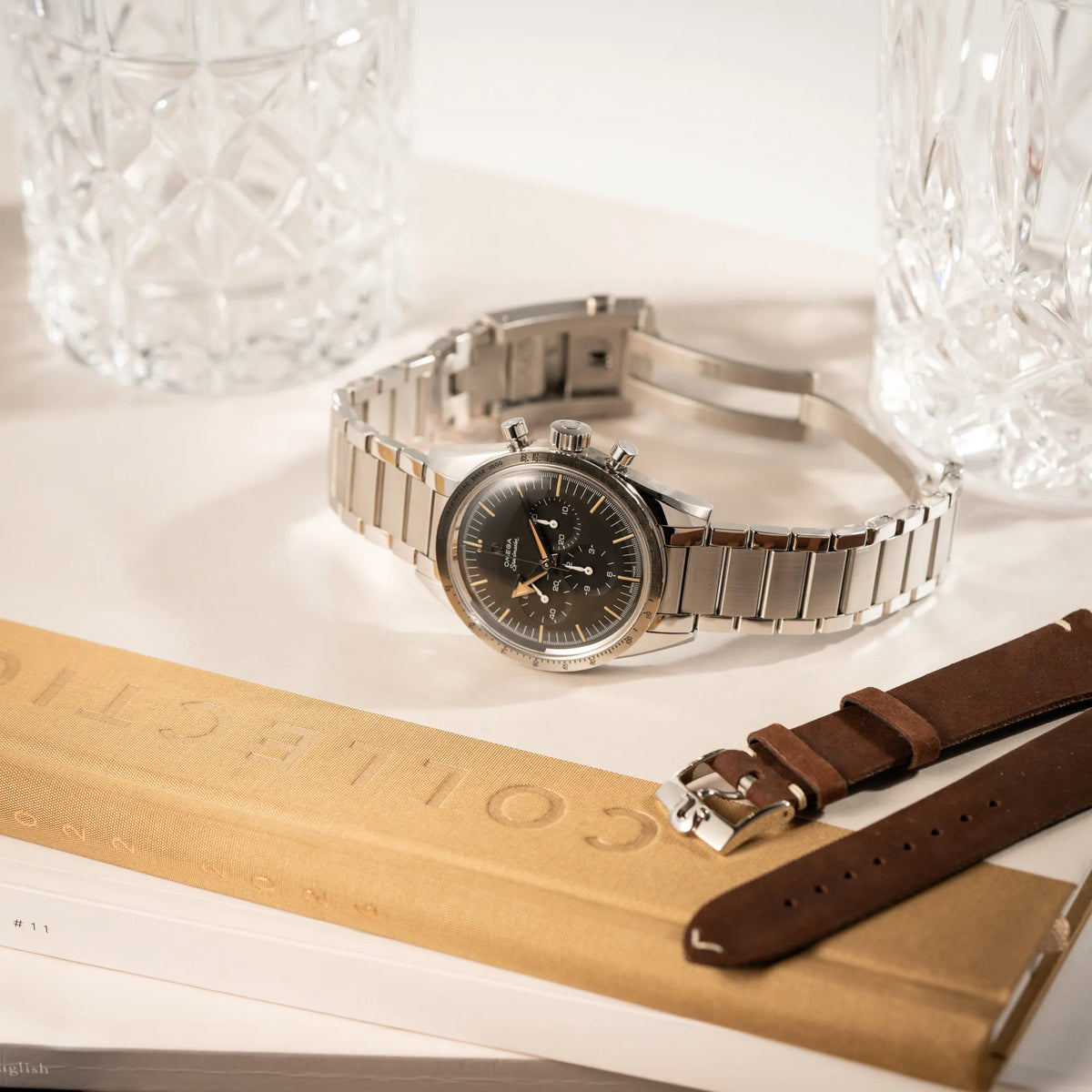 Produktfotografie der Omega Speedmaster 1957 Trilogy Limited Edition mit einem anthrazitfarbenen Zifferblatt mit Vintage-Look und dem original Omega-Stahlband im Ed-White-Design zusammen mit dem zusätzlichen Omega-Lederband in braun mit originaler Dornschliesse