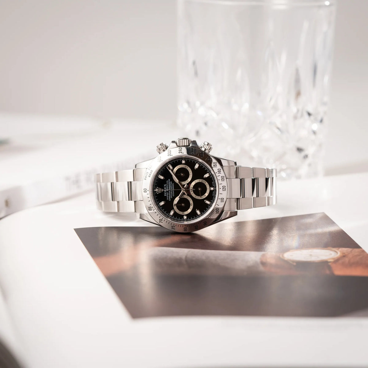 Produktfotografie der auf einem Uhrenbuch liegenden Rolex Cosmograph Daytona 116520 mit schwarzem Zifferblatt und der klassischen Edelstahl-Lünette vor Umstellung auf die Keramik-Lünette