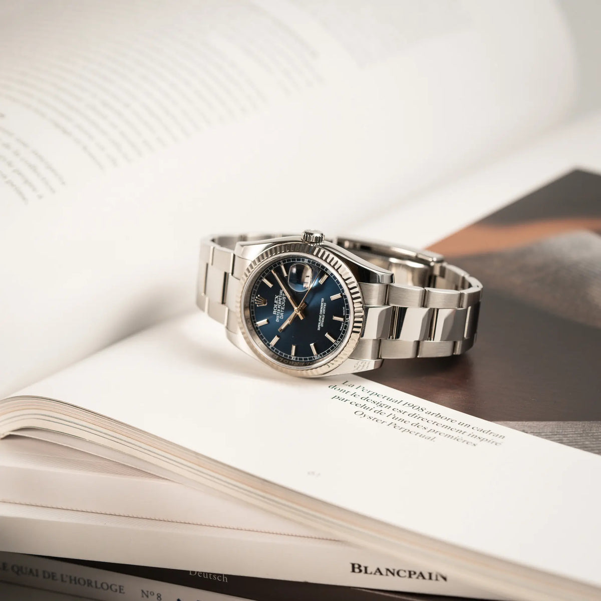 Produktfotografie der Rolex Datejust 36 der Referenz 116234 mit geriffelter Lünette und blauem Sonnenschliff-Zifferblatt während die Uhr auf einem Uhrenbuch liegt
