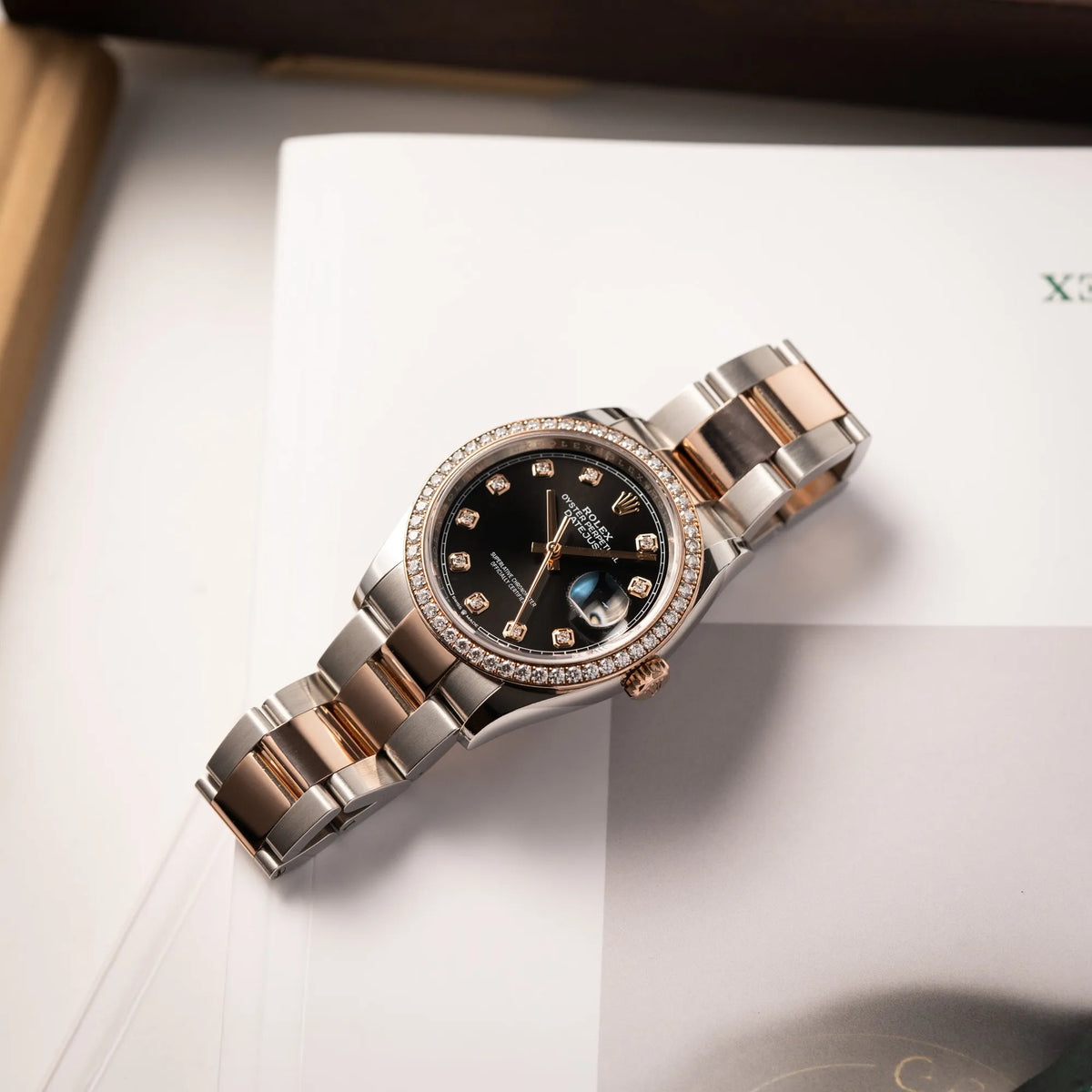 Produktfotografie der flach auf einem Rolex-Buch liegenden Rolex Datejust 36 126218RBR in Stahl-Roségold mit schwarzem Diamant-Zifferblatt und Diamant-Lünette im Factory-Besatz