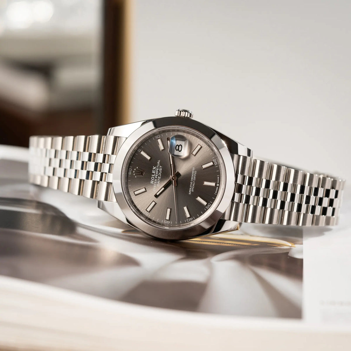 Produktfotografie der seitlich auf dem Uhrengehäuse liegenden Rolex Datejust 41 der Referenz 126300 mit grauem Zifferblatt und glatter Lünette am Jubilee-Stahl-Armband
