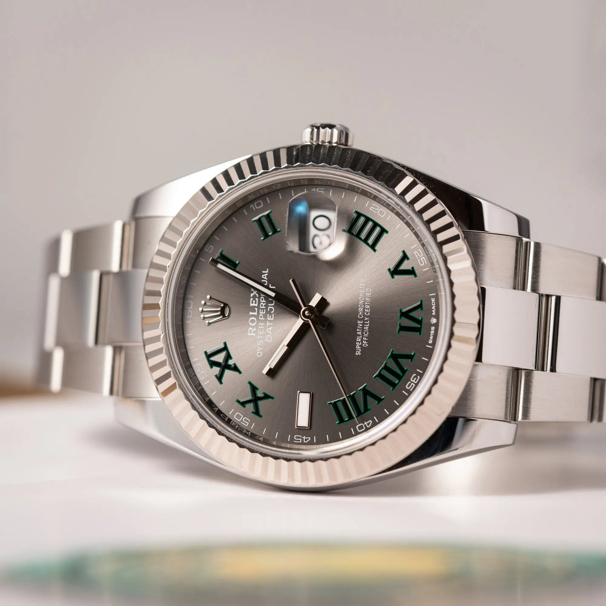 Produktfotografie der auf dem seitlichen Uhrengehäuse liegenden Rolex Datejust 41 der Referenz 126334 mit geriffelter Weißgold-Lünette und Oyster-Stahlband mit dem grauen Wimbledon-Zifferblatt mit schwarz-grünen Indizes