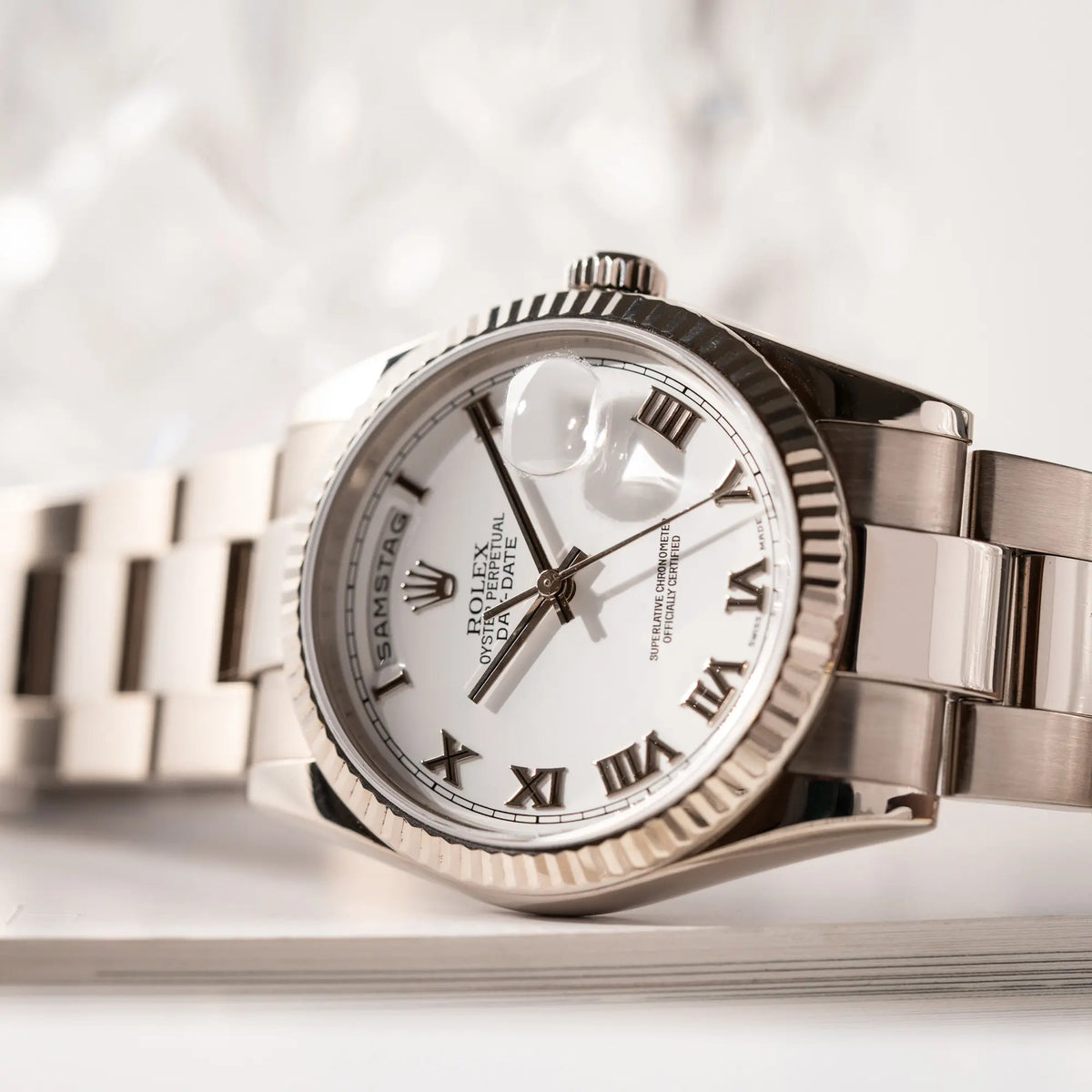 Produktfotografie der Rolex Day-Date 36 Armbanduhr in Weißgold der Referenz 118239 mit weißem Zifferblatt mit römischen Indizes am Oyster-Weißgold-Armband zeigt den Uhrenkopf, während die Uhr seitlich auf der Gheäuseflanke liegt