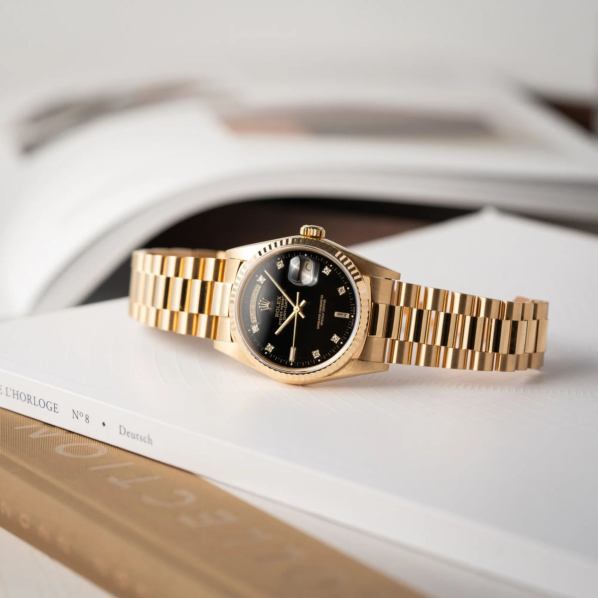 Produktfotografie der Rolex Day-Date 36 der Referenz 18238 in Gelbgold mit schwarzen Diamond-Dial am President-Armband, während die Uhr auf einem Buch liegt