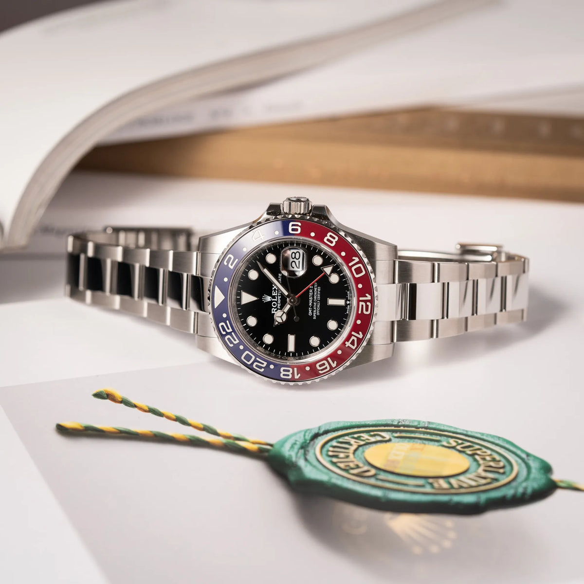 Produktfotografie der Rolex GMT-Master II der Referenz 126710BLRO mit der blau-roten Pepsi-Lünette in Keramik am Oyster-Stahl-Armband im ungetragenen Zustand, während die Uhr auf einem Buch liegt, welches ein Rolex Hang-Tag abbildet