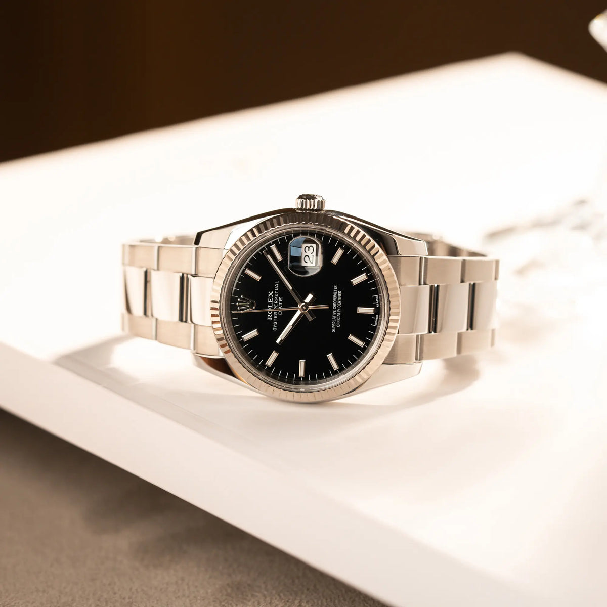 Produktfotografie der seitlich auf dem Uhrengehäuse liegenden Rolex Oyster Perpetual Date 34 der Referenz 115234 mit schwarzem Zifferblatt und der geriffelten Weißgold-Lünette 
