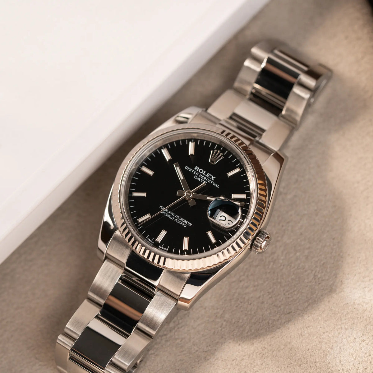 Rolex Oyster Perpetual Date 34 der Referenz 115234 mit schwarzem Zifferblatt und der geriffelten Weißgold-Lünette  liegt auf einem Juwelierstablet