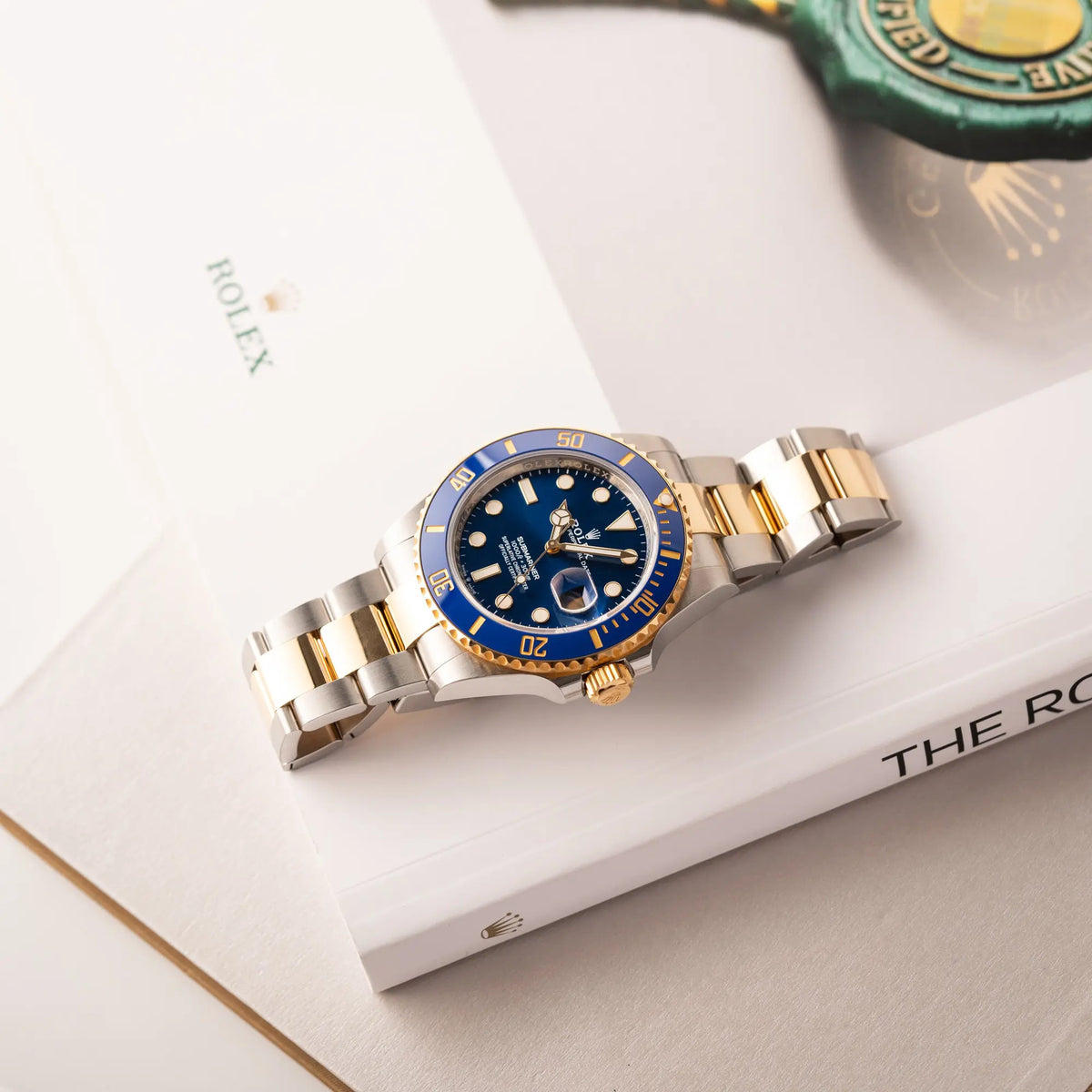 Produktfotografie zeigt die Rolex Submariner Date 126613LB Bluesy mit der typischen Kombination aus blauem Zifferblatt und Lünette mit dem Gehäuse und Armband aus Stahl-Gold flach auf einem Uhrenbuch liegend