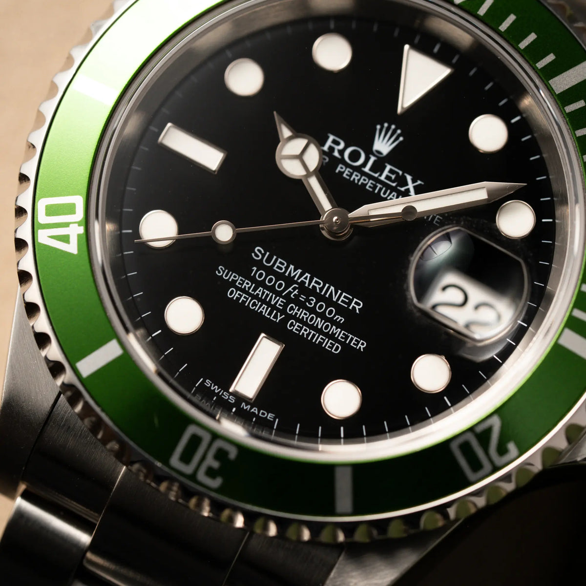 Makroaufnahme der Rolex Submariner Date 16610LV aus 2007 mit schwarzem Zifferblatt und der grünen "Kermit" Aluminium-Lünetten-Einlage mit Fokus auf dem Zeigersatz und dem Submariner Schriftzug bei 6 Uhr