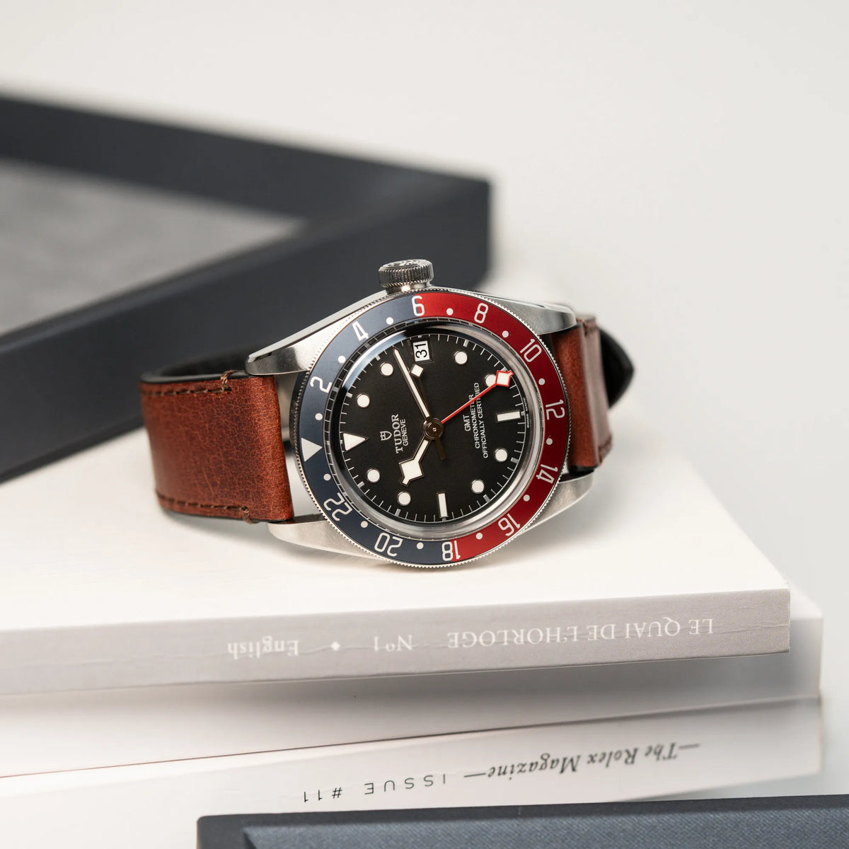 Produktfotografie der Tudor Black Bay Pepsi mit der Referenz 97830RB am braunen Lederband im Neuzustand, während die Uhr auf einem Horologie Buch liegt