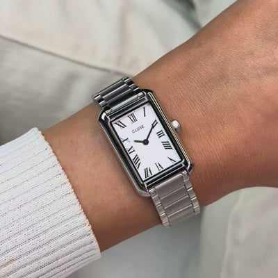Frau trägt die silberfarbene Cluse Belisenna Damenuhr mit länglichem,  rechteckigem Gehäuse und weißem Zifferblatt mit römischen Indizes an ihrem Handgelenk und präsentiert die Uhr in die Kamera