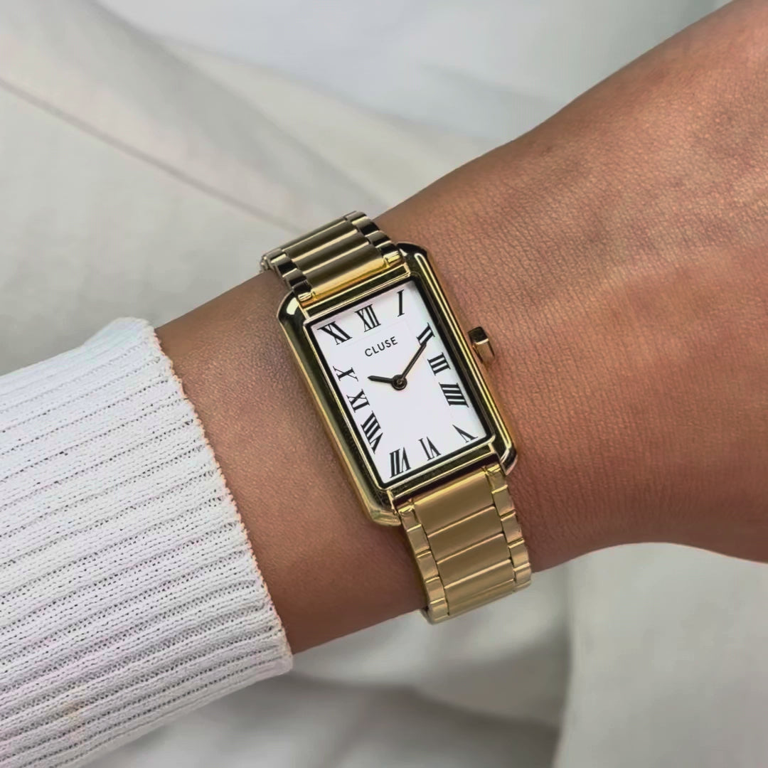 Frau trägt die goldfarbene Cluse Belisenna Damenuhr mit länglichem,  rechteckigem Gehäuse und weißem Zifferblatt mit römischen Indizes an ihrem Handgelenk und präsentiert die Uhr in die Kamera