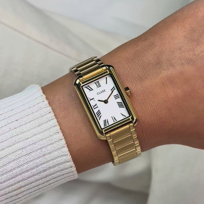 Frau trägt die goldfarbene Cluse Belisenna Damenuhr mit länglichem,  rechteckigem Gehäuse und weißem Zifferblatt mit römischen Indizes an ihrem Handgelenk und präsentiert die Uhr in die Kamera